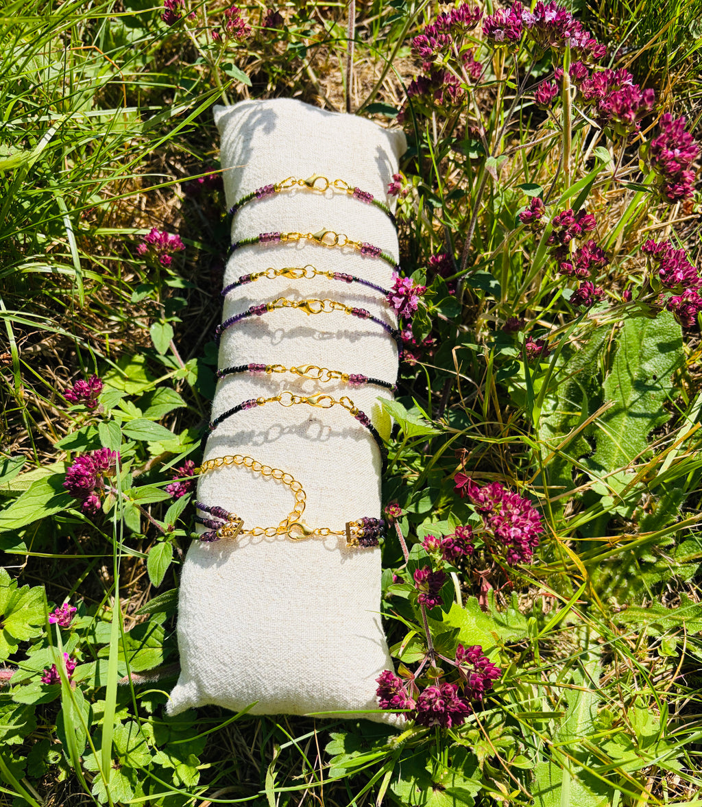 A set of gold chain bracelets with dark purple beads is displayed on a white fabric jewelry pillow, placed outdoors on grass and surrounded by purple wildflowers.