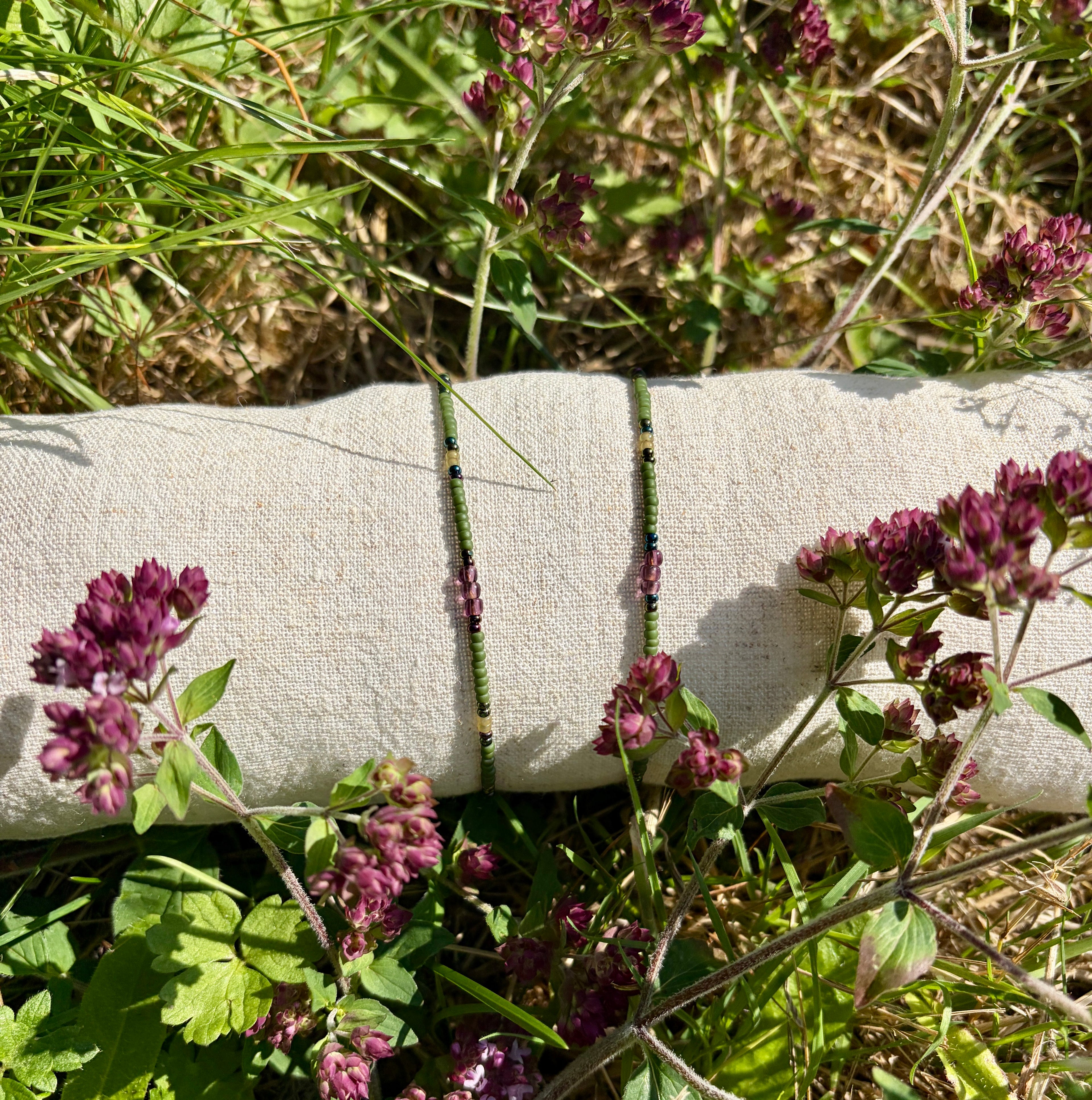 Two beaded bracelets with purple flower charms and green beads, displayed on a rolled beige fabric sleeve outdoors among green leaves and purple flowers.