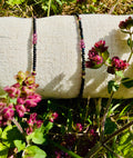 Two matte black beaded bracelets on a linen pillow sitting in purple wildflowers